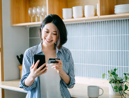 A woman is standing in a modern kitchen holding a smartphone in one hand and a credit card in the other, suggesting an online payment or mobile banking activity.