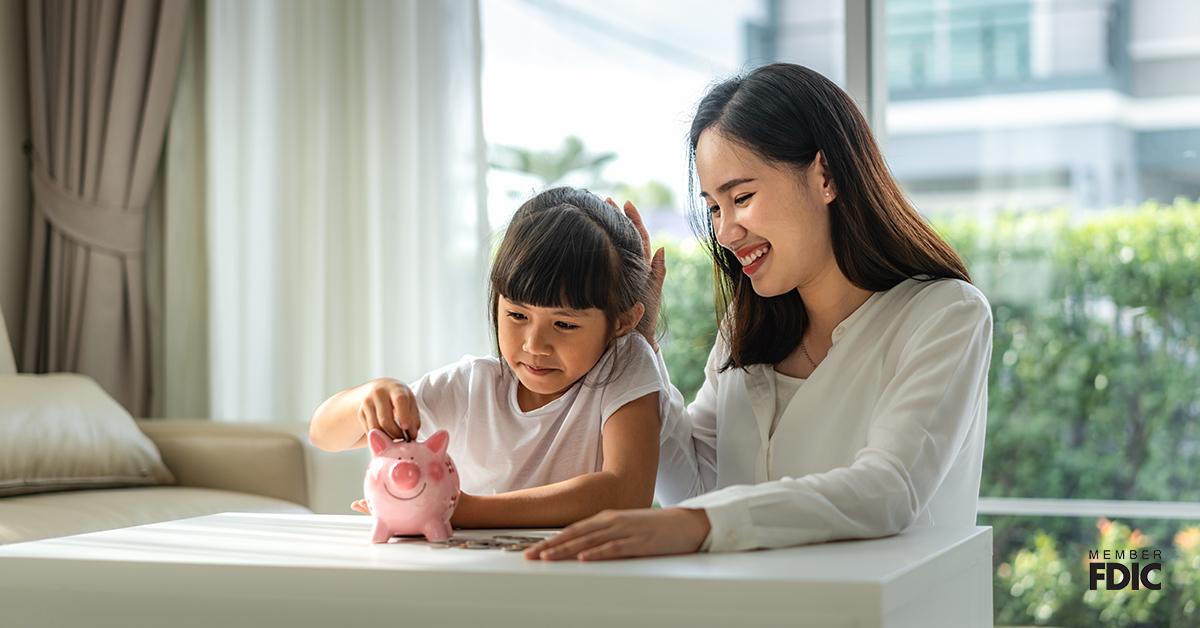 Happy mother and daughter saving money putting coins into piggy bank on table at home.