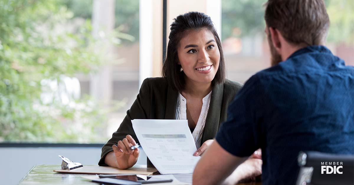 A cheerful female banker sits at her desk while speaking to a client about easy account transfers. 