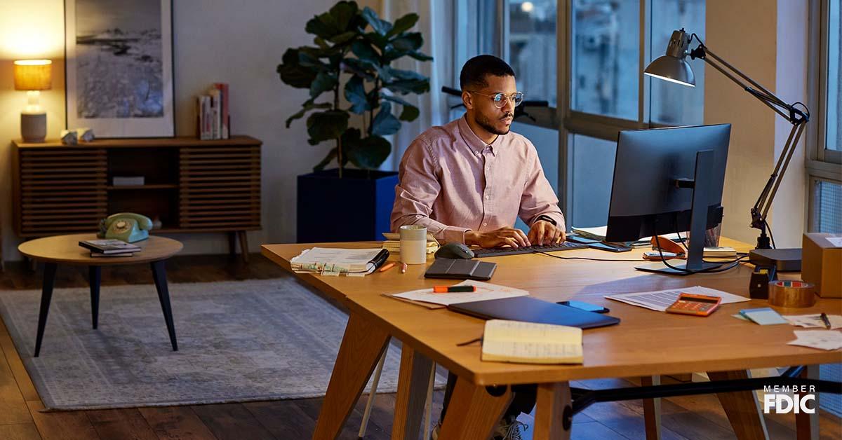 A man sits with glasses sits at his work from home desk in the evening, focusing on the computer screen. 