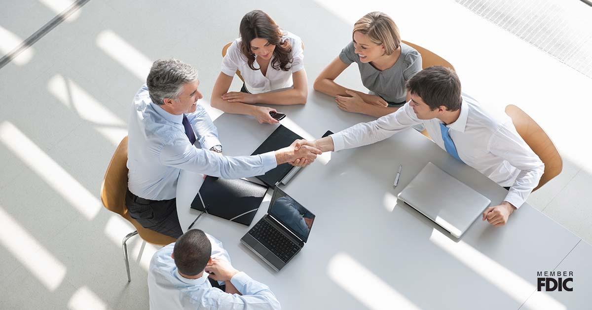 A group of professionals are gathered around a conference table shaking hands as they make a business deal. 