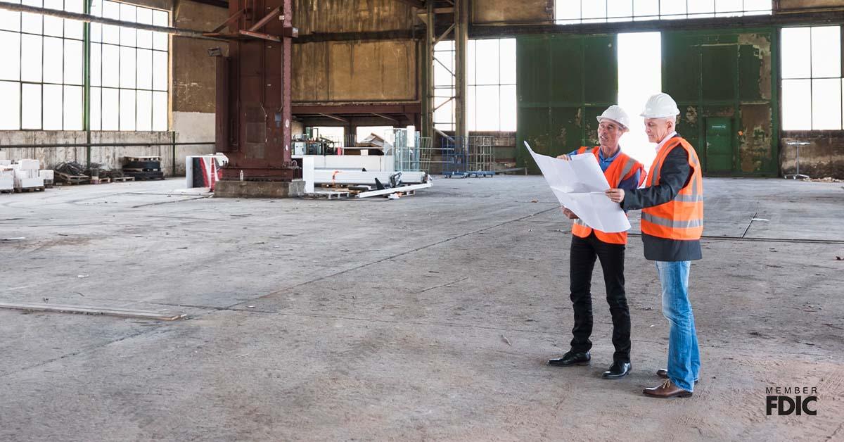 Dos hombres con chalecos naranjas y cascos blancos se encuentran dentro de un gran almacén con papeles de construcción para su próxima empresa. 