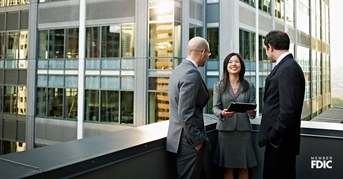 Three professionals are networking outside of a business building while the female professional smiles at her two male colleagues holding a digital tablet.