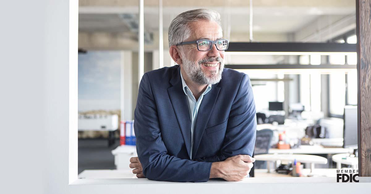 A middle-aged man with glasses and grey hair smiles as he leans over a window inside his small business office. 