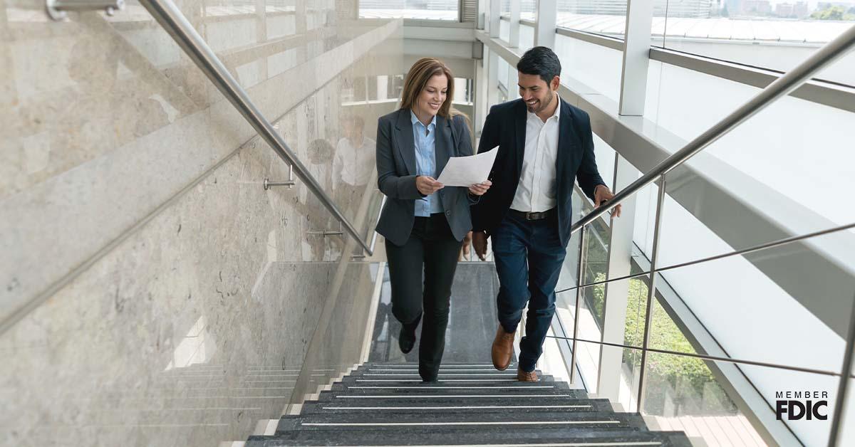一位女商人和她的男同事在一个开放空间办公室走上楼梯，同时查看一份企业信贷额度文件。