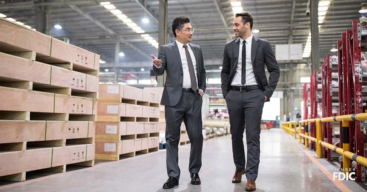Two business men walk together through a warehouse to discuss cross-border financing.