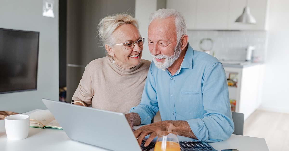 A middle-aged couple sits at a kitchen table while smiling at each other and looking at their wealth management finances on a laptop.