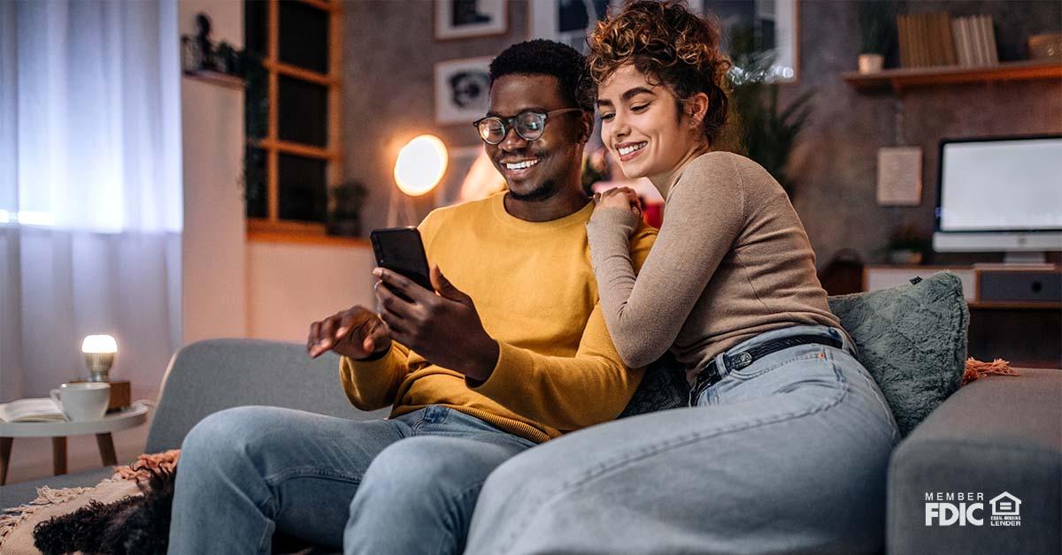 Una pareja joven se sienta en un sofá de la sala de estar y sonríe a su teléfono móvil mientras revisa sus opciones de hipoteca de Cathay Bank.