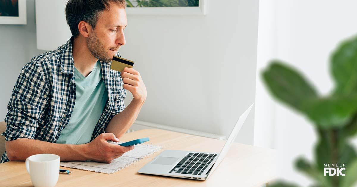 Un hombre se sienta en su escritorio de madera en una oficina bien iluminada mientras mira su computadora portátil y sostiene su tarjeta de crédito con expresión preocupada por las alertas de fraude.