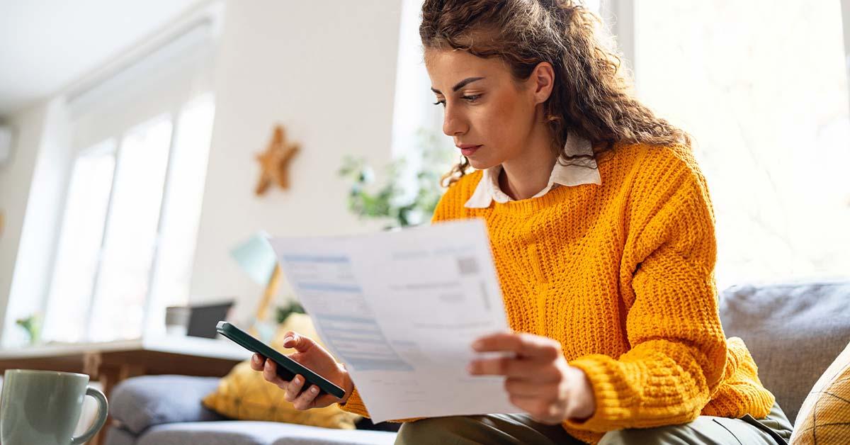 A woman wearing a yellow sweater looks cozy while sitting on the couch in her living room while holding important financial documents and checking her credit history on her cell phone.