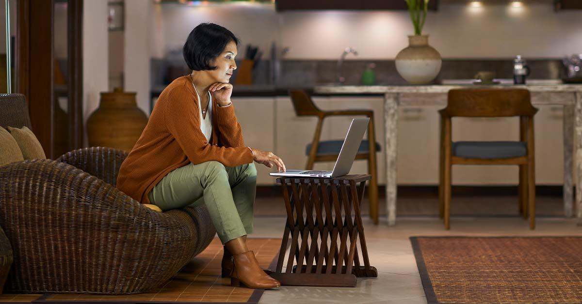 A woman with short black hair sits on a couch at home while trying to open an online banking account using her laptop.
