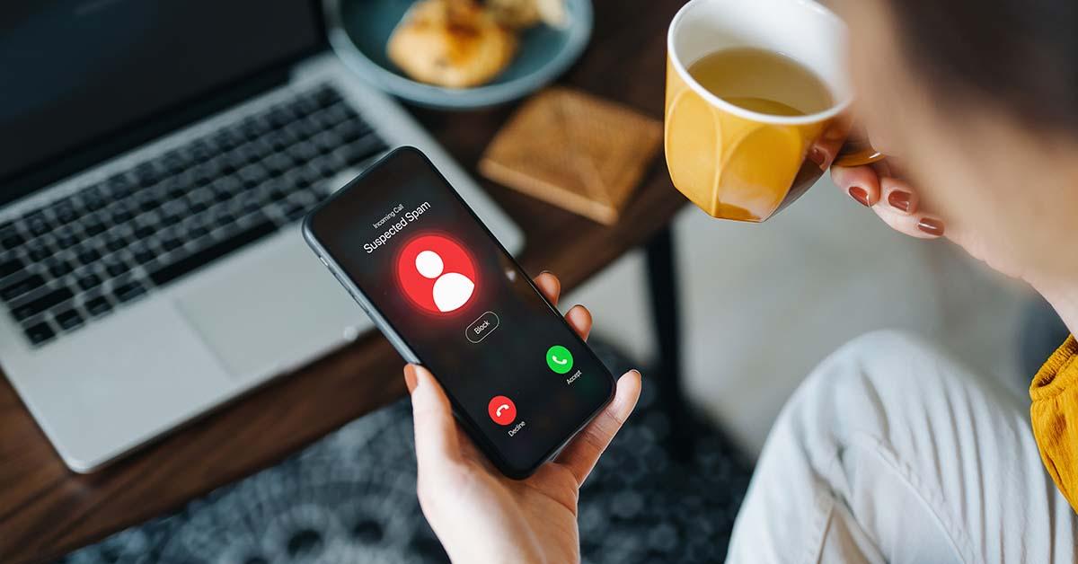 Over the shoulder view of young Asian woman receiving an incoming suspected call from unknown caller on her smartphone and rejecting the call at home. Device screen showing warning sign as detected by the network provider.