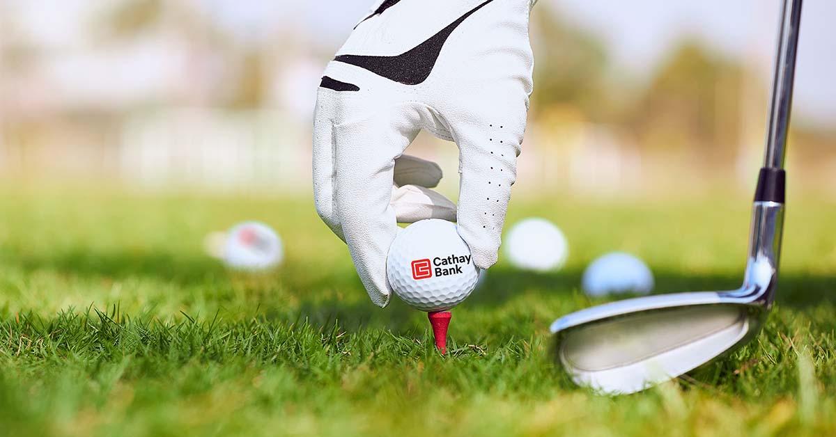 A Cathay Bank Golf Ball sits on the grass at an outdoor golf course with a golf club behind it.