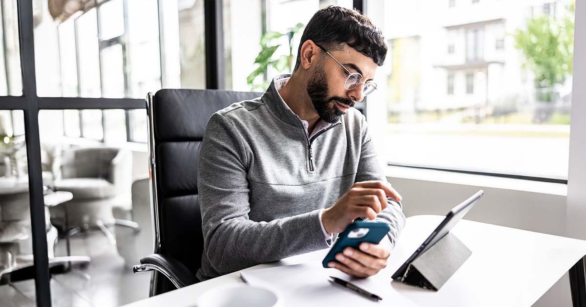 一位身穿灰色四分之一拉链毛衣、戴着眼镜的男子坐在工作桌前。他左手拿着手机,右手则专注地指向桌上摆着的 iPad。