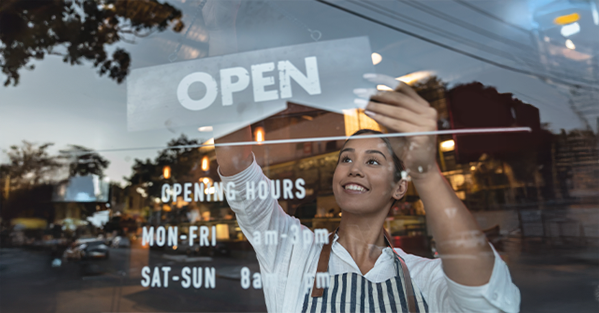 一位小企业主正在调整商店橱窗上的“营业中”标志。橱窗上倒映着街道、汽车和温暖的室内灯光，展现出一个充满活力的社区景象。