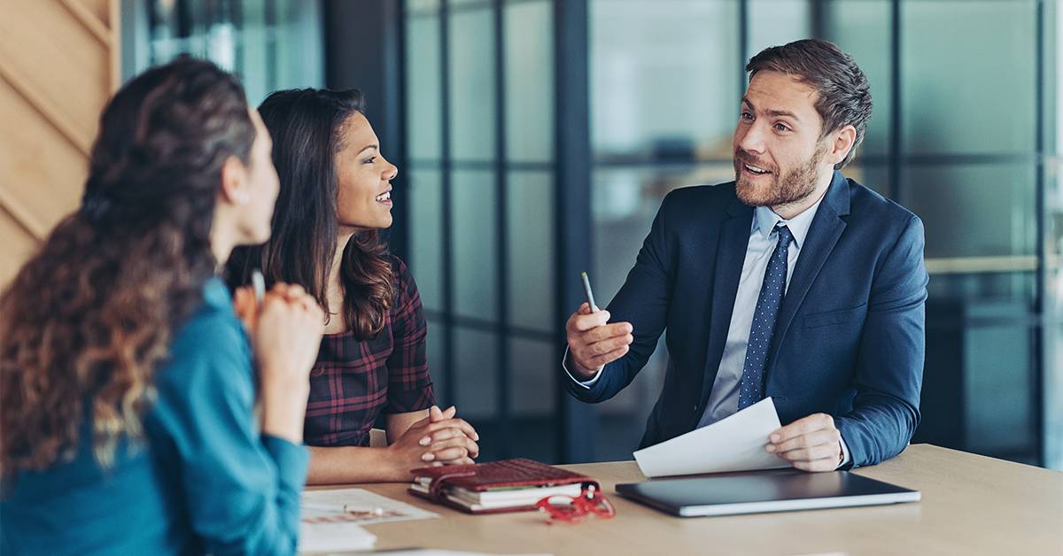Three professionals in a modern office discuss advanced banking strategies, with documents, a notebook, and a laptop on the table during a collaborative meeting.