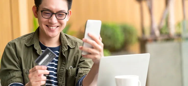  A man is sitting at a table outdoors with a laptop, holding a smartphone in one hand and a credit card in the other, suggesting an online payment or mobile banking activity.