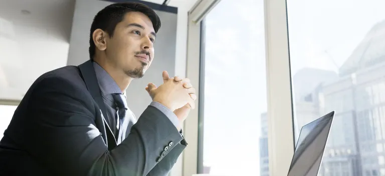 A businessman at laptop looking out urban office window