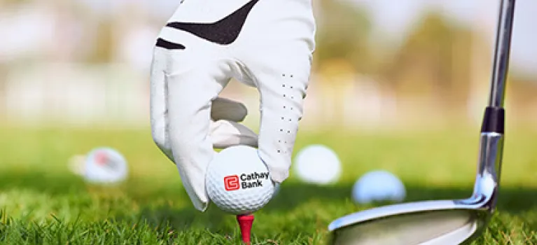 A golf ball with Cathay Bank logo sits outdoors on grass with a golf club and hand next to it for the annual tournament in California. 