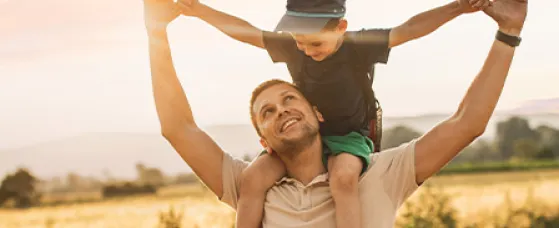 A young boy sits atop his father’s shoulders while walking down the road near a field with mountains in the background. 