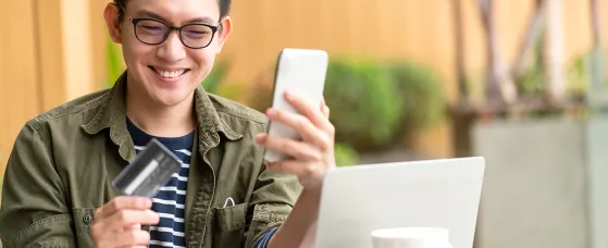  A man is sitting at a table outdoors with a laptop, holding a smartphone in one hand and a credit card in the other, suggesting an online payment or mobile banking activity.