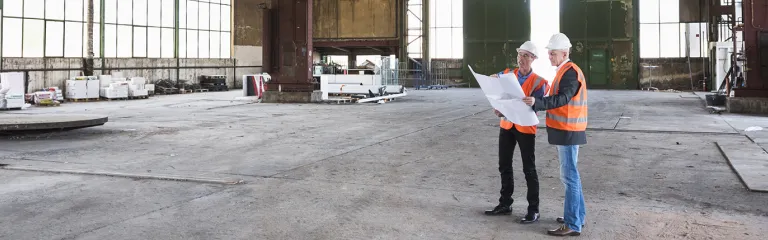 Two men with plan wearing safety vests talking in old industrial hall