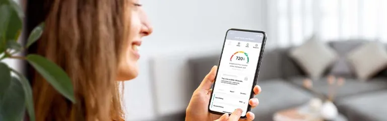 A woman smiles as she checks her credit score on a mobile phone while sitting in a furnished room with a grey couch and matching pillows.