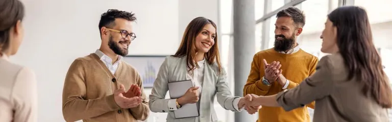 Four professionals in a modern office shake hands during a meeting while two others applaud. One person in a light suit holds a tablet, and the group stands near a table with charts in the background.