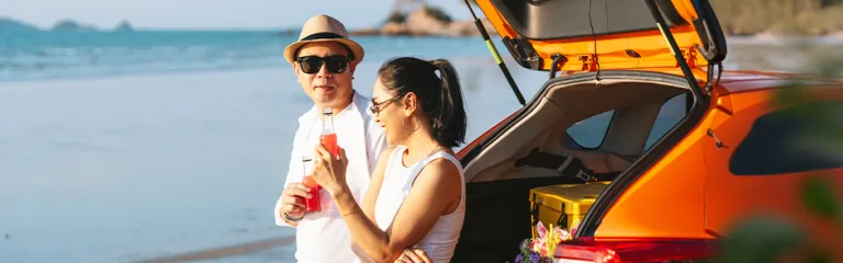 Two people relaxing by an orange SUV parked on a beach, holding cold drinks and enjoying a summer day by the ocean.